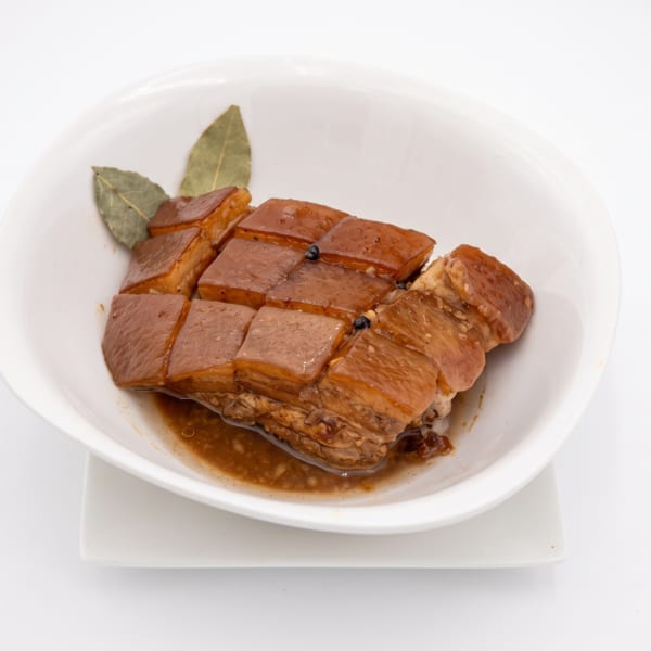 Braised pork belly, cut into squares, in a white bowl with bay leaves and peppercorns.