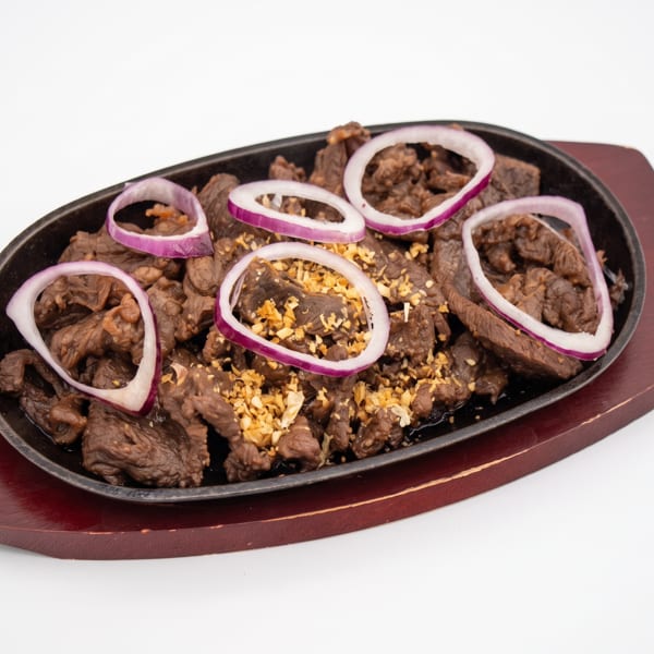 Sizzling beef salpicao with garlic bits and red onion rings on a black platter.